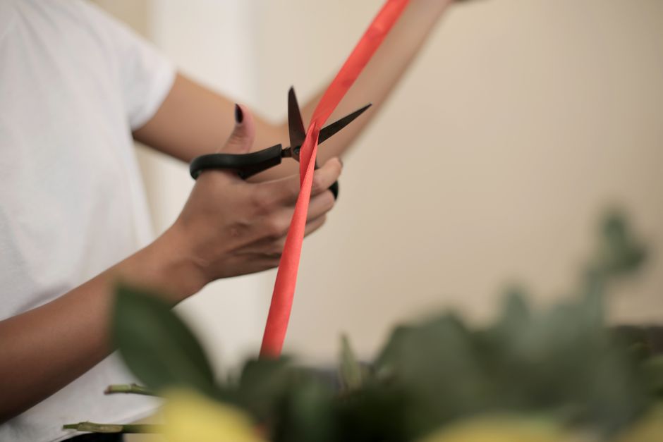 Close-up of a person’s hands holding a pair of black scissors and trimming a coral-colored ribbon around a floral bouquet. The individual is wearing a white short-sleeved shirt, and the background f