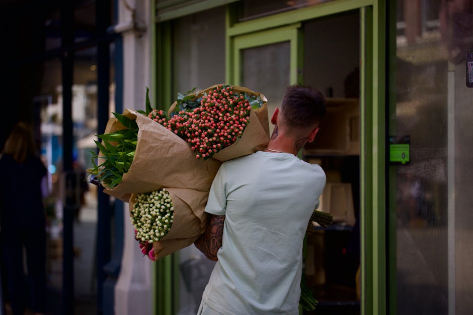 A man with short dark hair and a sleeve tattoo on his right arm, wearing a light-colored T-shirt, is seen from behind holding a large bouquet of fresh flowers wrapped in brown kraft paper. The bouquet