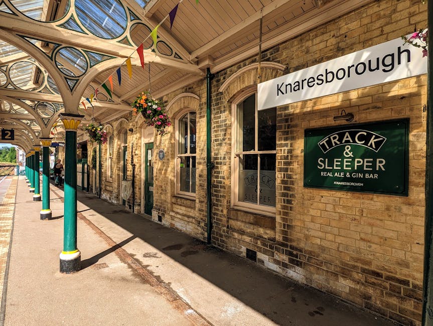 Photograph of Knaresborough station platform with a covered waiting area featuring decorative white arched supports and hanging flower baskets with pink and purple blossoms. The brick station building