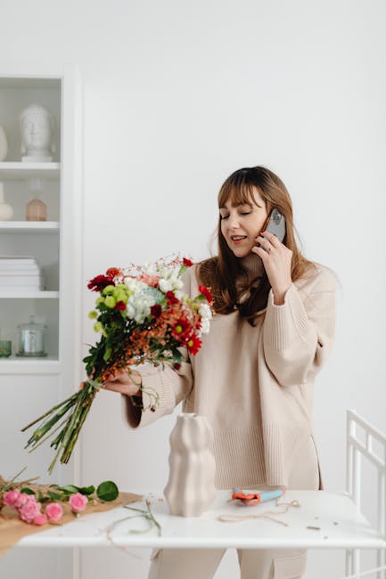 A woman with long brown hair and bangs, dressed in a light beige sweater, is placing a large, vibrant floral bouquet into a white vase on a white table. The bouquet features a variety of fresh flowers