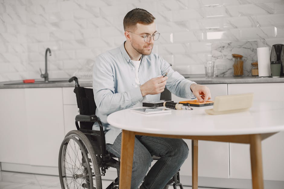 A young man with short brown hair, glasses, and light skin, seated in a wheelchair at a white round table in a modern kitchen, is carefully holding a small yellow flower, surrounded by a bouquet of fr