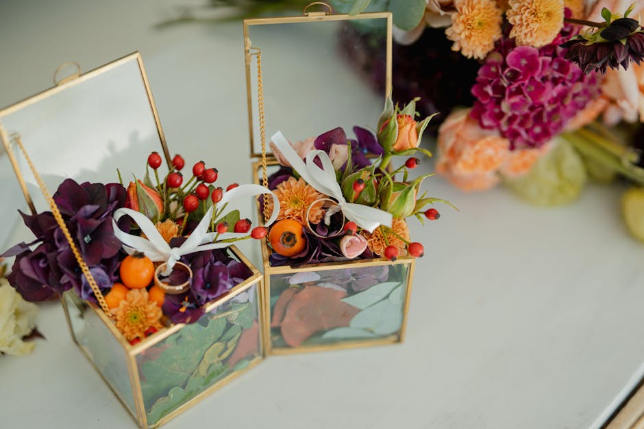Two small glass and gold photo frame boxes are used as floral gift containers, each filled with an arrangement of fresh flowers. One box contains dark purple calla lilies, orange ranunculus, red berri