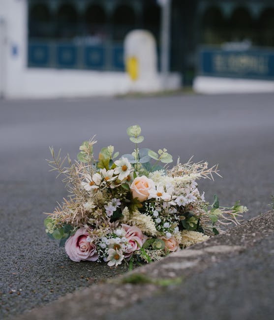 A delicate bouquet of fresh flowers lying on the pavement near a curb, arranged with pale pink roses, white daisies, and small clusters of baby's breath, complemented by greenery and wispy, dried foli