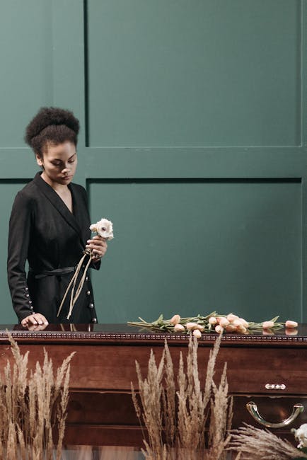 A young woman with dark curly hair styled in an updo, wearing a black blazer, stands in front of a green wall during a floral arrangement event. She holds a small bouquet of white and pale pink flower
