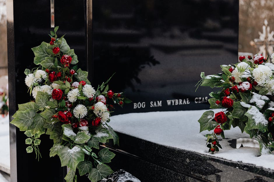A black polished gravestone decorated with fresh flower arrangements featuring red roses, white chrysanthemums, and green variegated ivy, with a light dusting of snow on the surface and floral bouquet