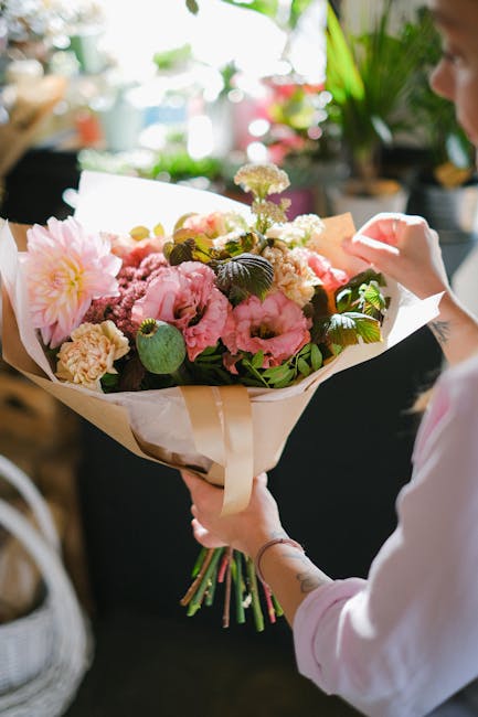 A person holding a large bouquet of fresh flowers, including pink peonies, white carnations, and greenery, wrapped in cream-colored paper with a satin ribbon. The arrangement is being presented indoor