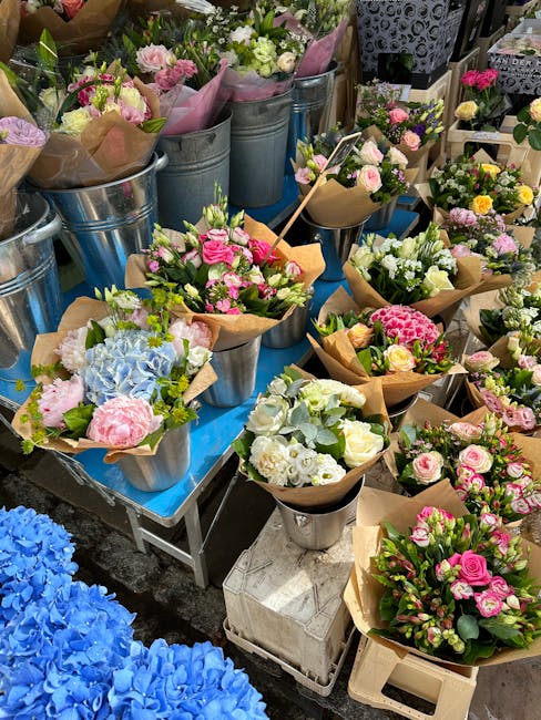 A display of fresh floral bouquets arranged in metal buckets and wrapped in brown craft paper, featuring a variety of spring flowers including pink roses, white roses, pale pink peonies, light blue hy
